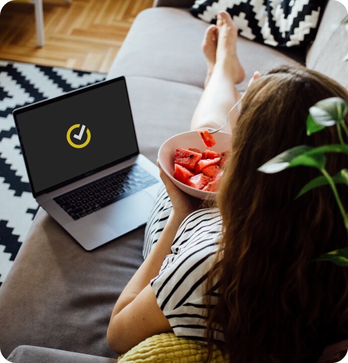 Mujer sobre un sofá con un tazón de fruta y un portátil a su lado con el logotipo de Norton